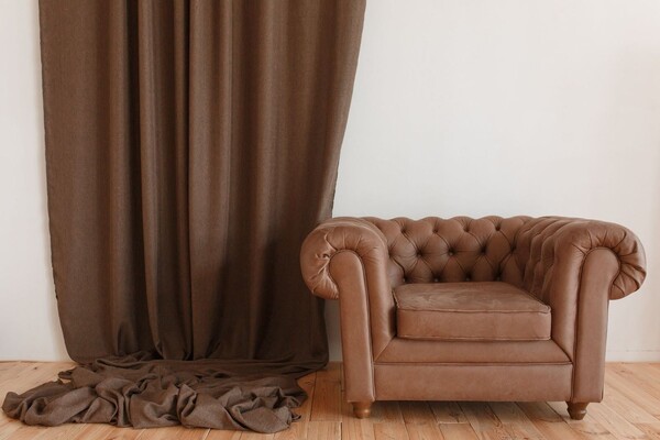 Klasyczny, skórzany fotel Chesterfield w ciemnym brązie, ustawiony obok grubego, płóciennego firana w podobnym kolorze, tworzy elegancki i spokojny klimat
